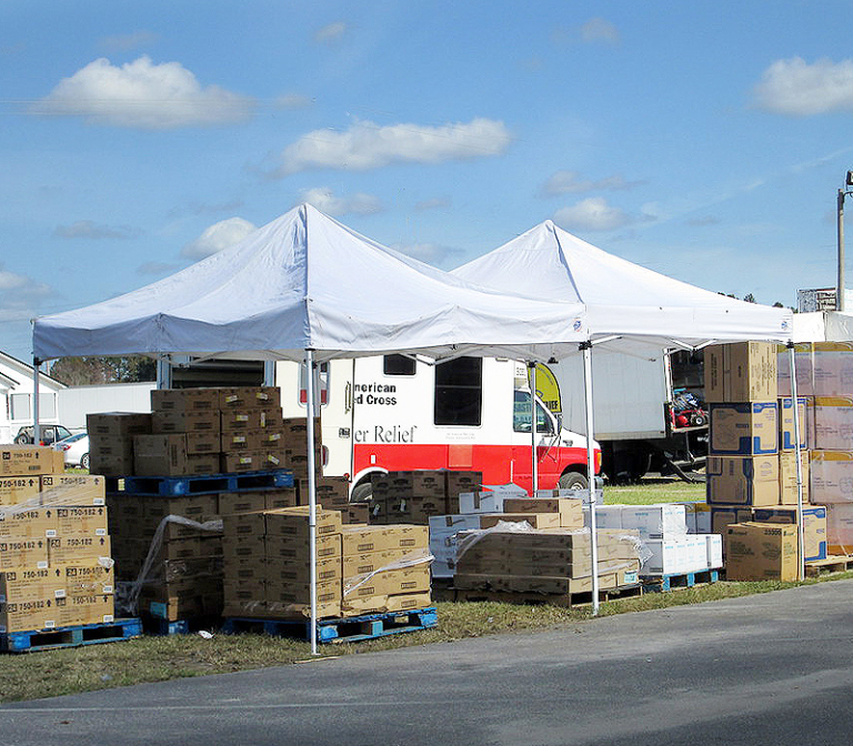 Two white pop up canopy tents shading pallets of boxed relief supplies beside a truck