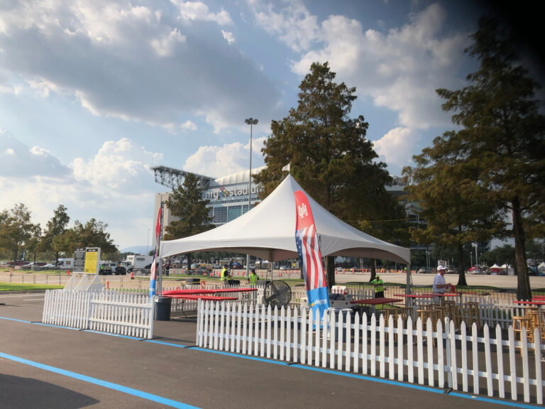 High peak marquee tent with red cocktail tables and a white picket fence in a stadium parking lot