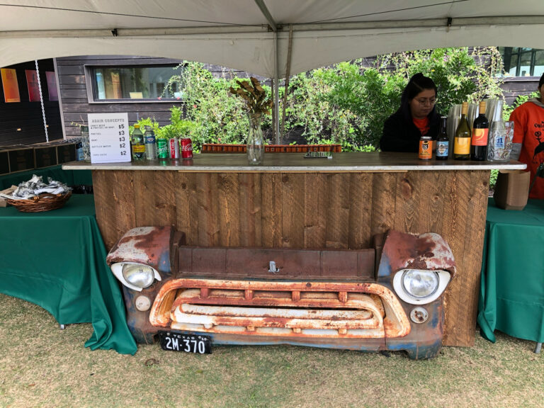 Rustic wood bar front built from a vintage truck grille under a tent, with bottled drinks and a menu sign