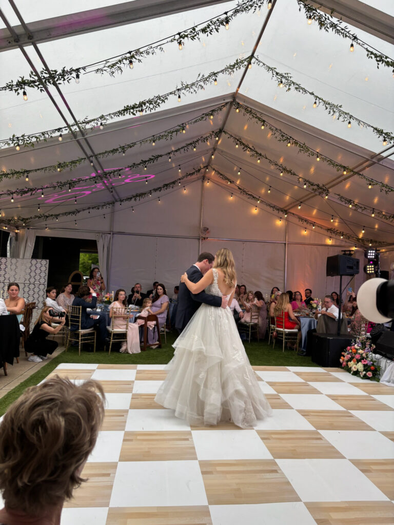A bride and groom dancing on a checkered dance floor under a clear top tent strung with lights and greenery